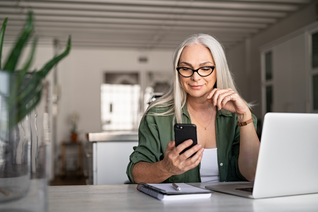 Happy senior woman using mobile phone while working at home with laptop